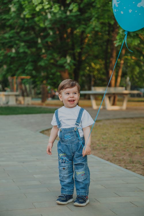 Content little child in overalls with balloon looking away on tiled walkway in urban park