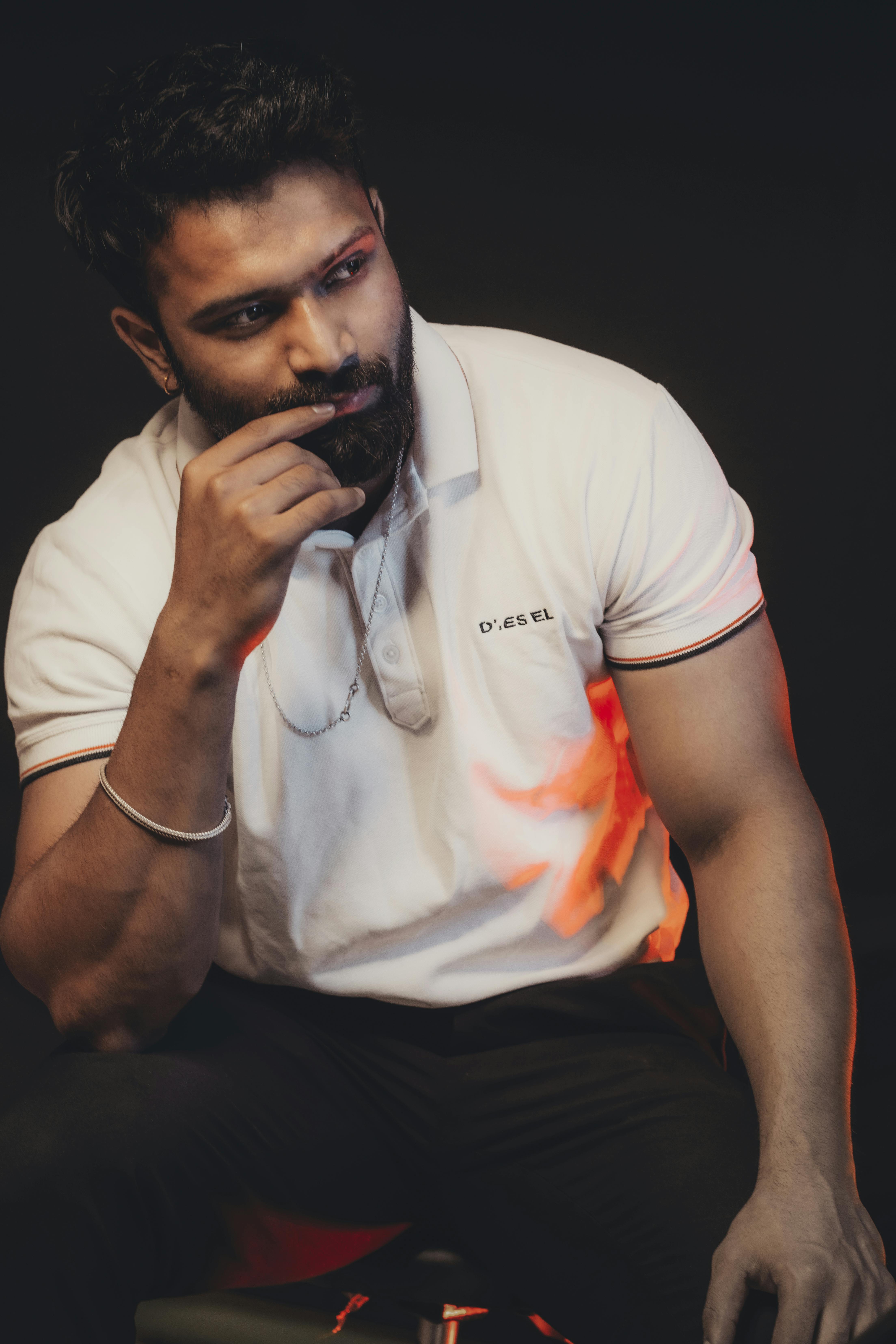 A bearded man in a white polo shirt poses pensively against a dark background.