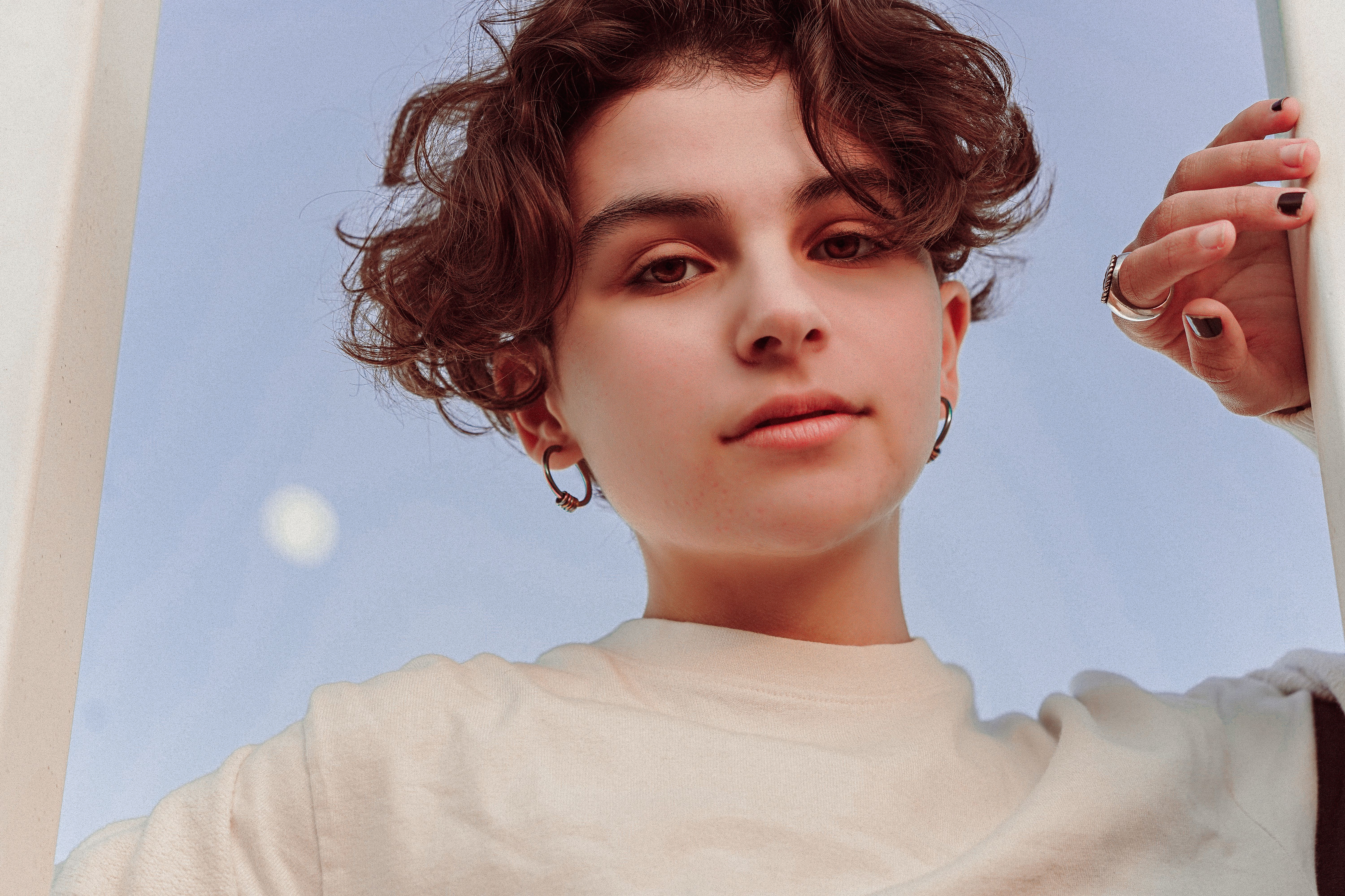 Close-up portrait of a young adult with curly hair, wearing earrings, under a clear sky.