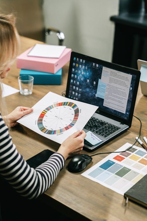 Free Businesswoman reviews color wheel and strategy documents on a laptop in an office setting. Stock Photo