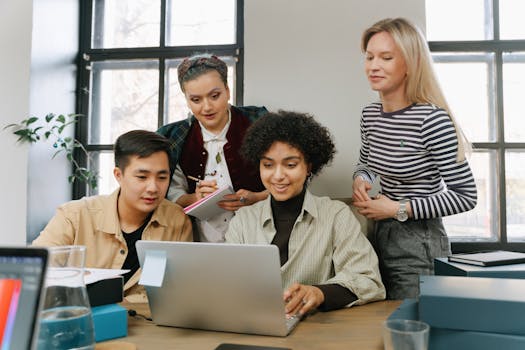 A diverse group of coworkers discuss strategies around a laptop in a bright office space.