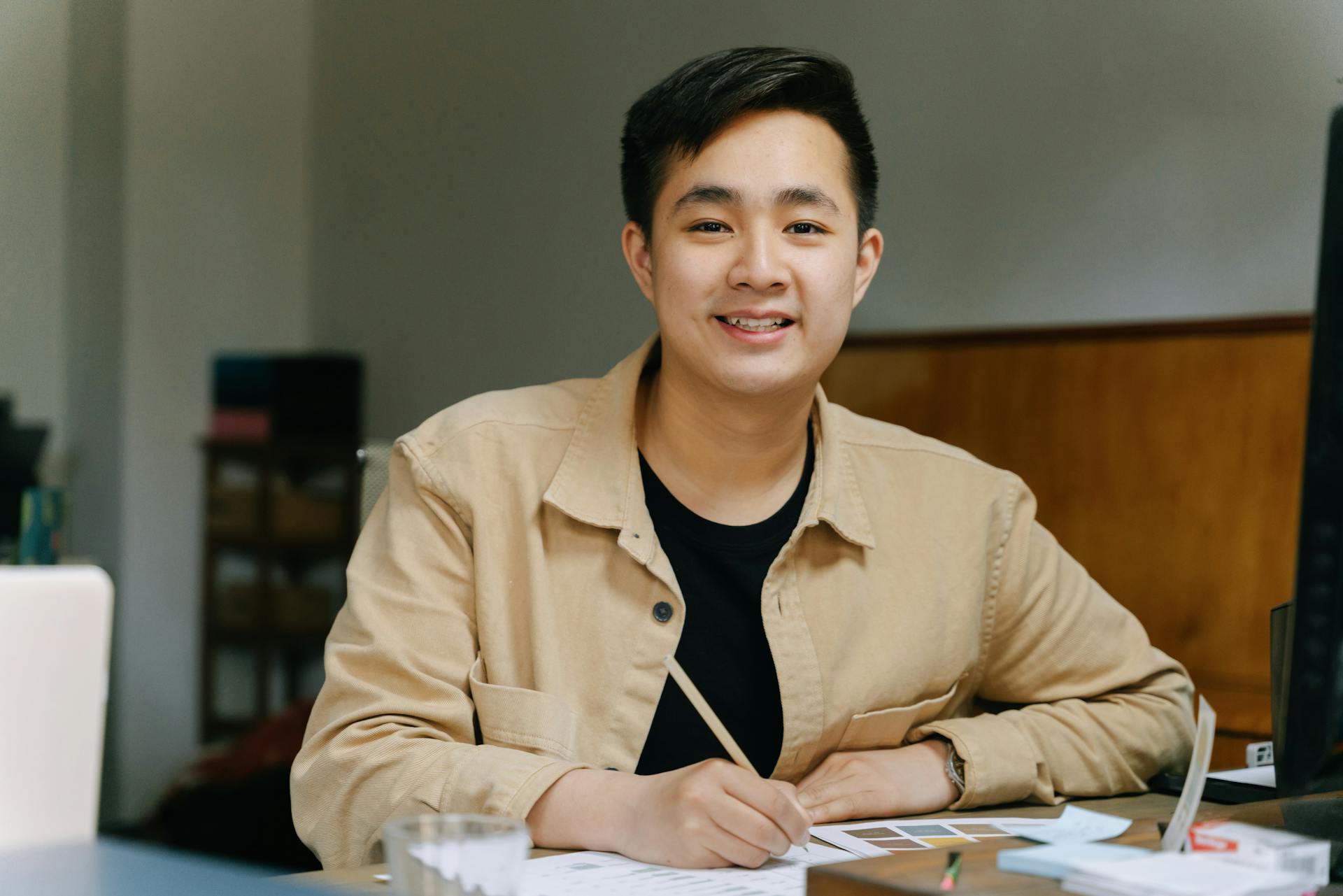 Asian man smiling and writing at a desk in a modern office setting.