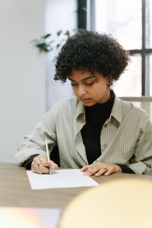 Free African American woman in a striped shirt writing on paper at a desk indoors. Stock Photo