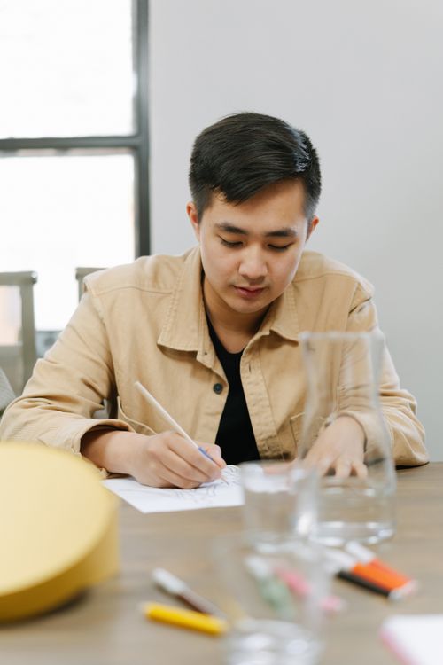 Free Young adult man in casual jacket writing at desk indoors with concentration. Stock Photo