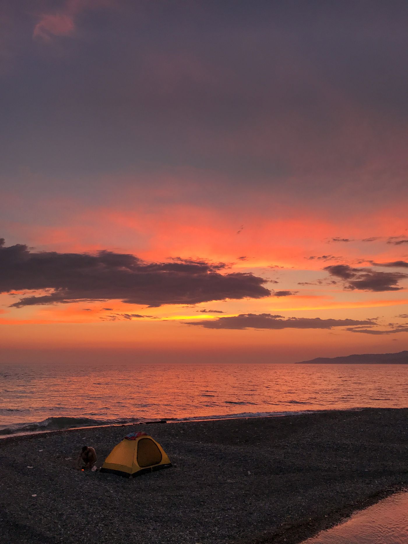 Person camping beside the beach with a bright tent at sunset