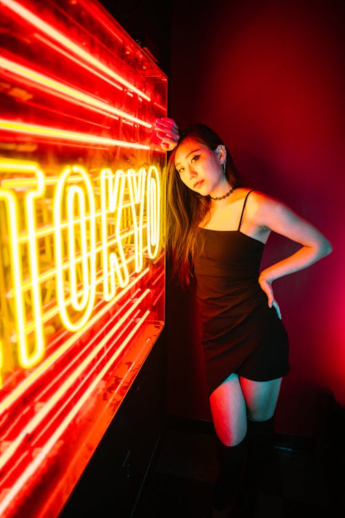 Woman in Black Spaghetti Strap Dress Leaning on Neon Signage