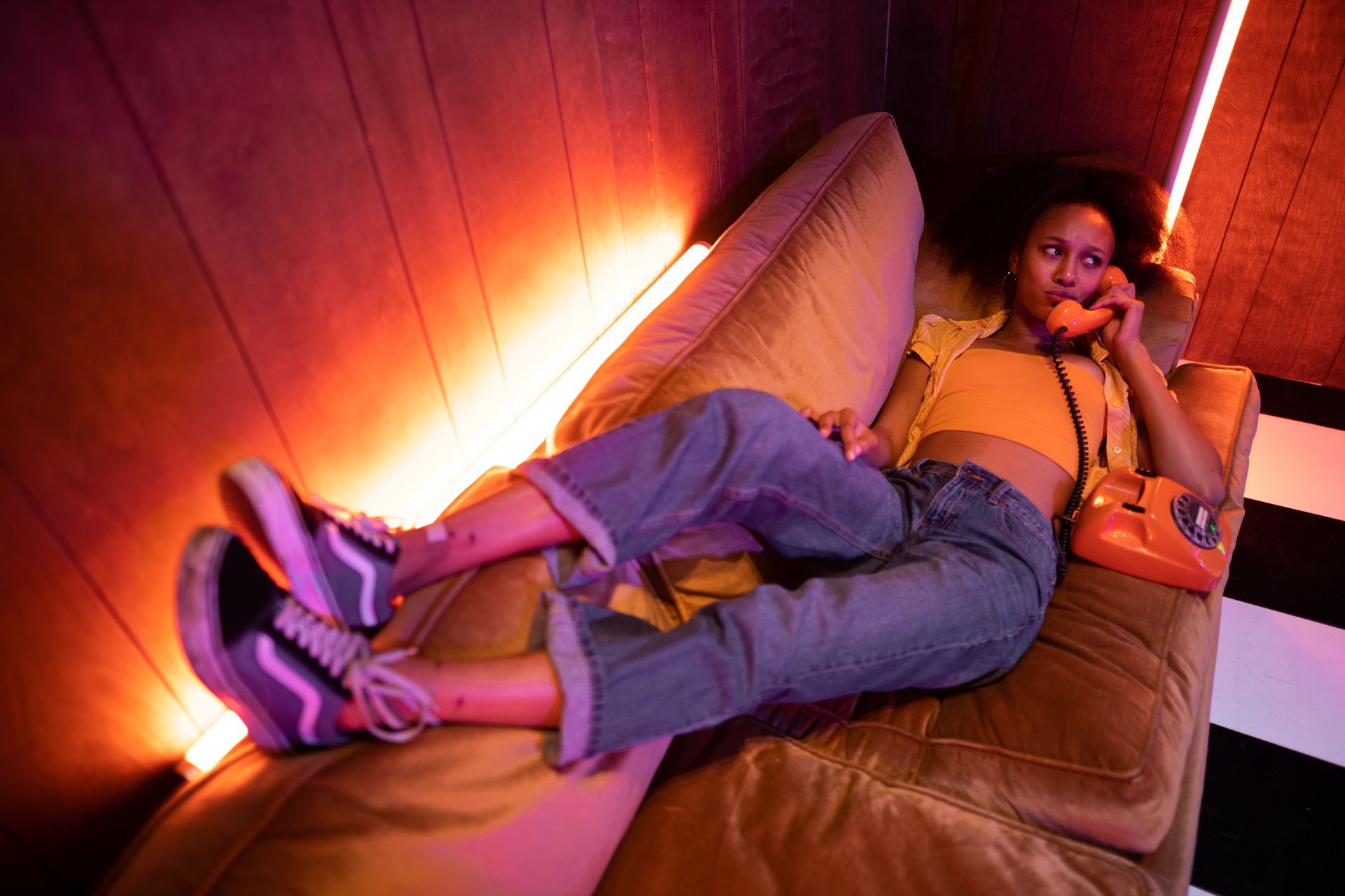A young woman lying on a sofa, talking on a vintage phone under neon lights.