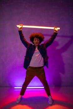 A stylish young man with curly hair holds a colorful neon light in a vibrant studio setting.
