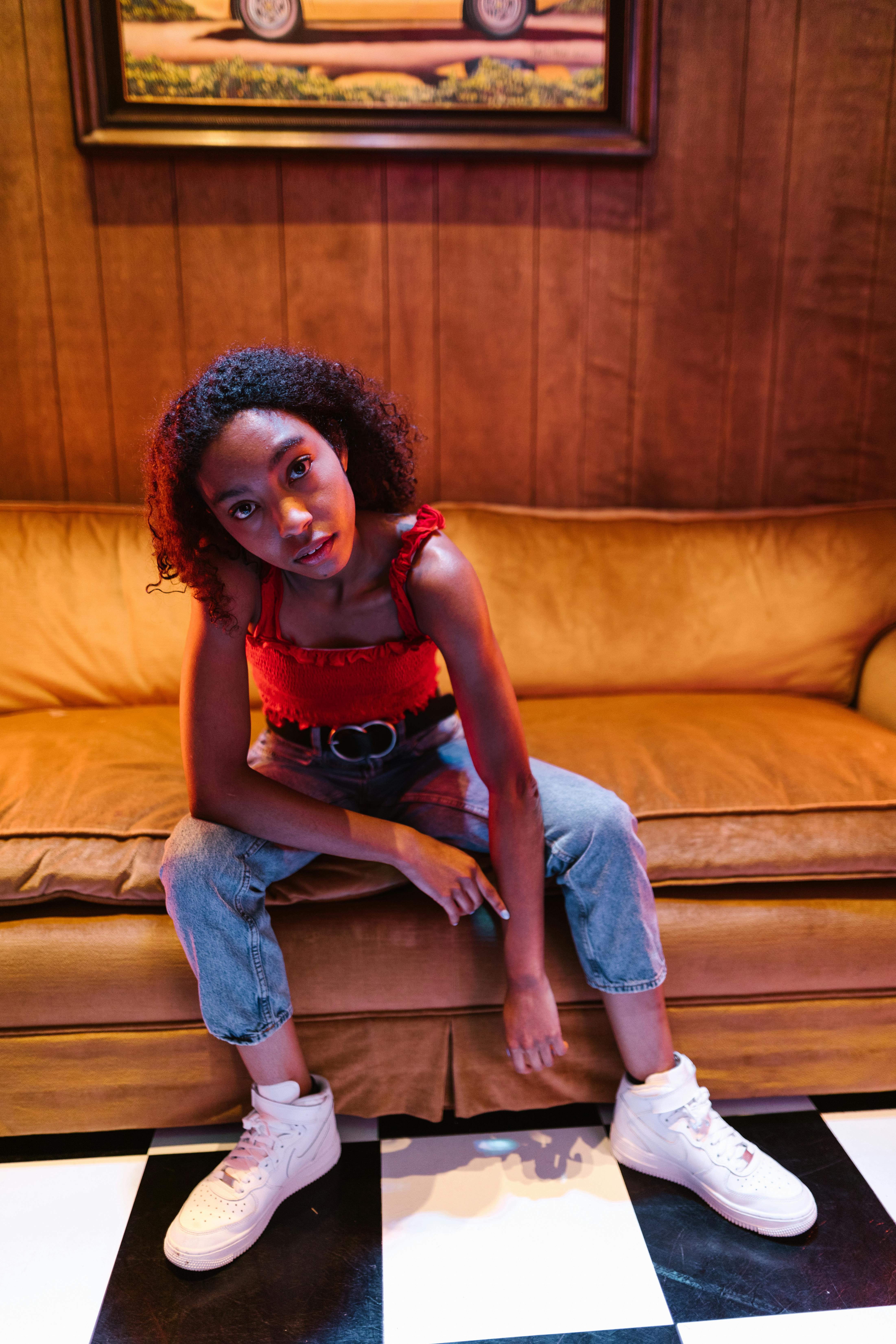 Fashionable young woman with afro hair posing on a couch in casual denim and sneakers.