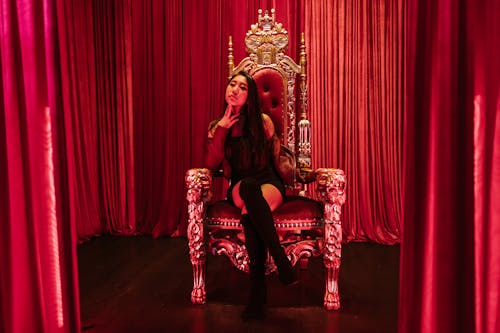 Glamorous woman in a black dress sitting on a decorative throne surrounded by red curtains.