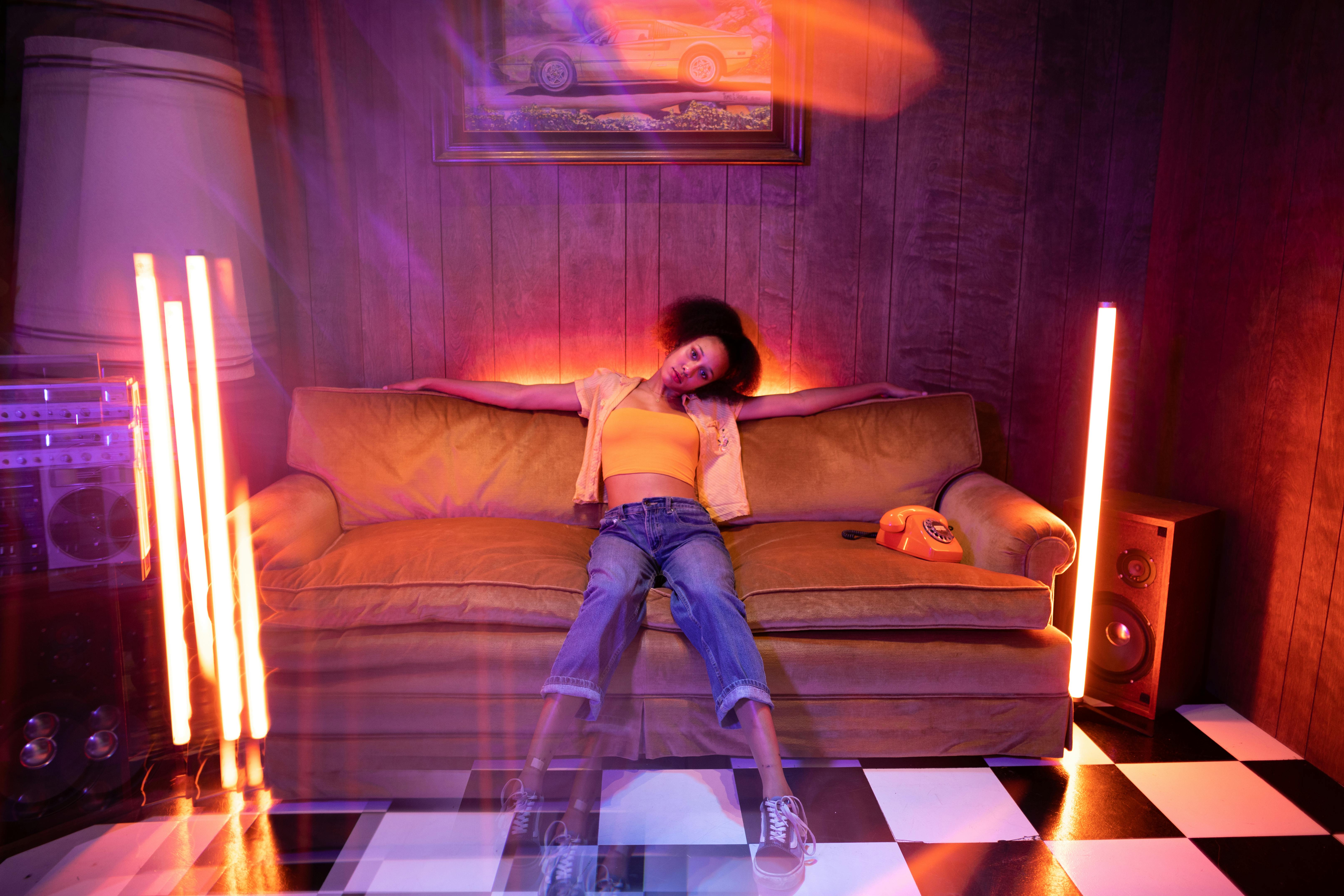 A young woman relaxes on a vintage couch with a vibrant retro setup, featuring neon lights and a classic telephone.