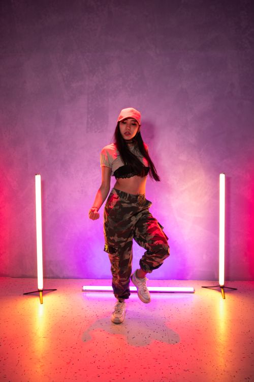 Free Young woman dancer in camouflage pants performing a hip-hop routine with vibrant neon lights creating a dynamic atmosphere. Stock Photo