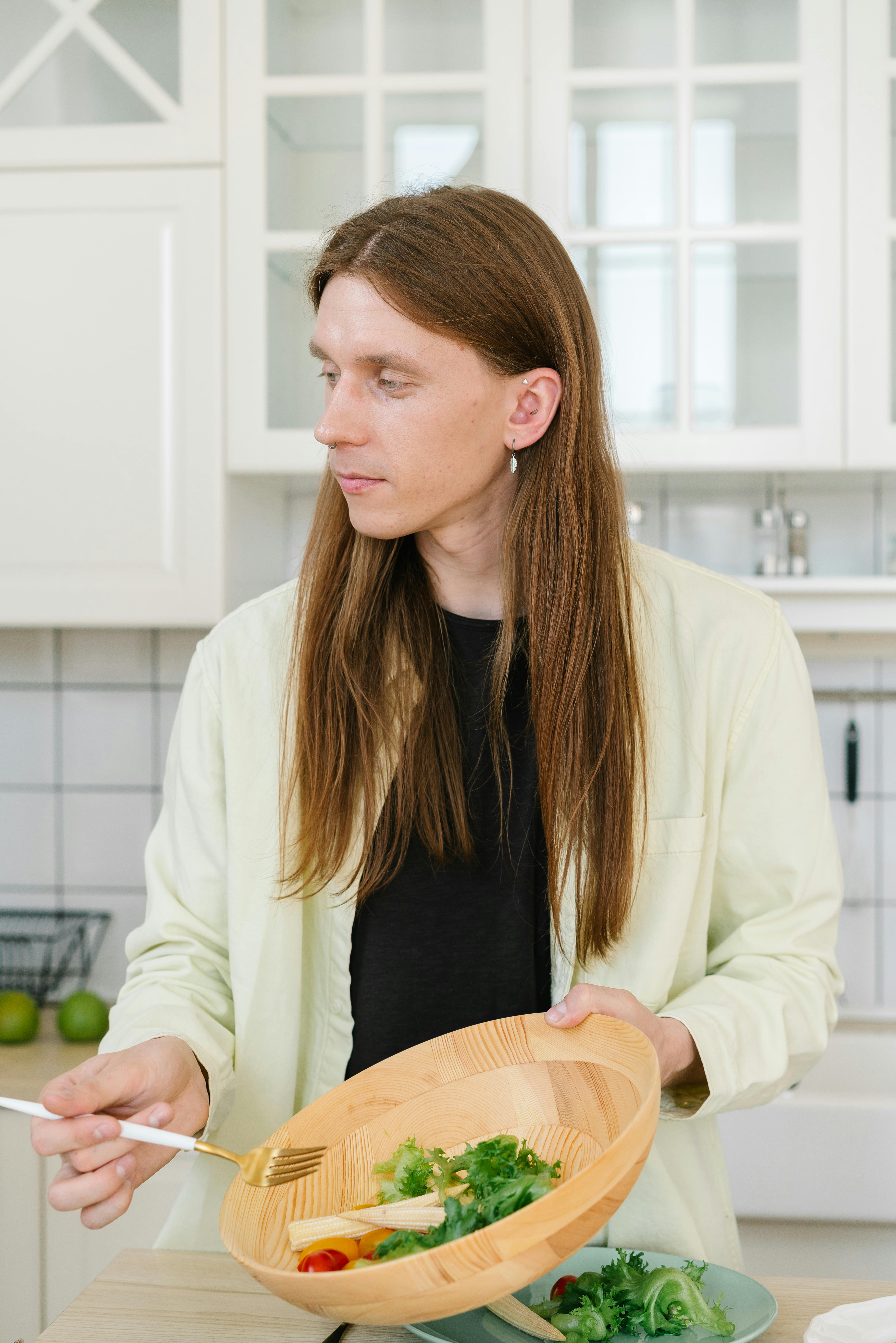 Young man with long hair holding wooden bowl in kitchen preparing a healthy salad.