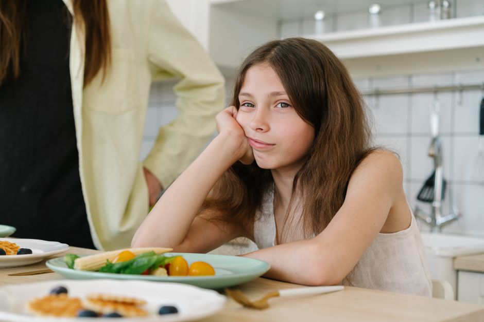 A young girl looking bored, sitting at a kitchen table with breakfast plates in front of her.