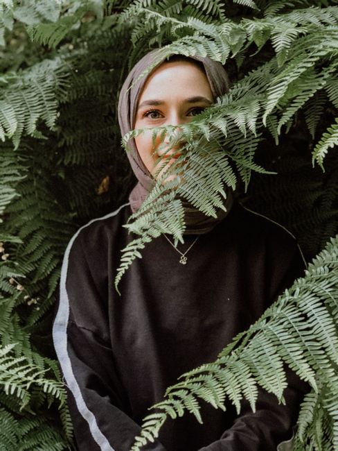 Young female in casual clothes and headscarf standing in garden with green plants while smiling and looking at camera
