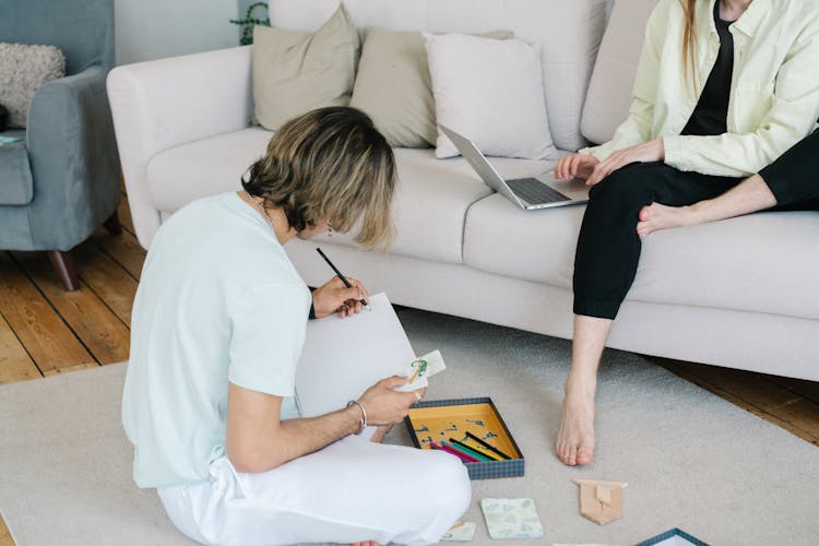 A Person Sitting On A Couch Working On A Laptop Near A Man Doing Artwork