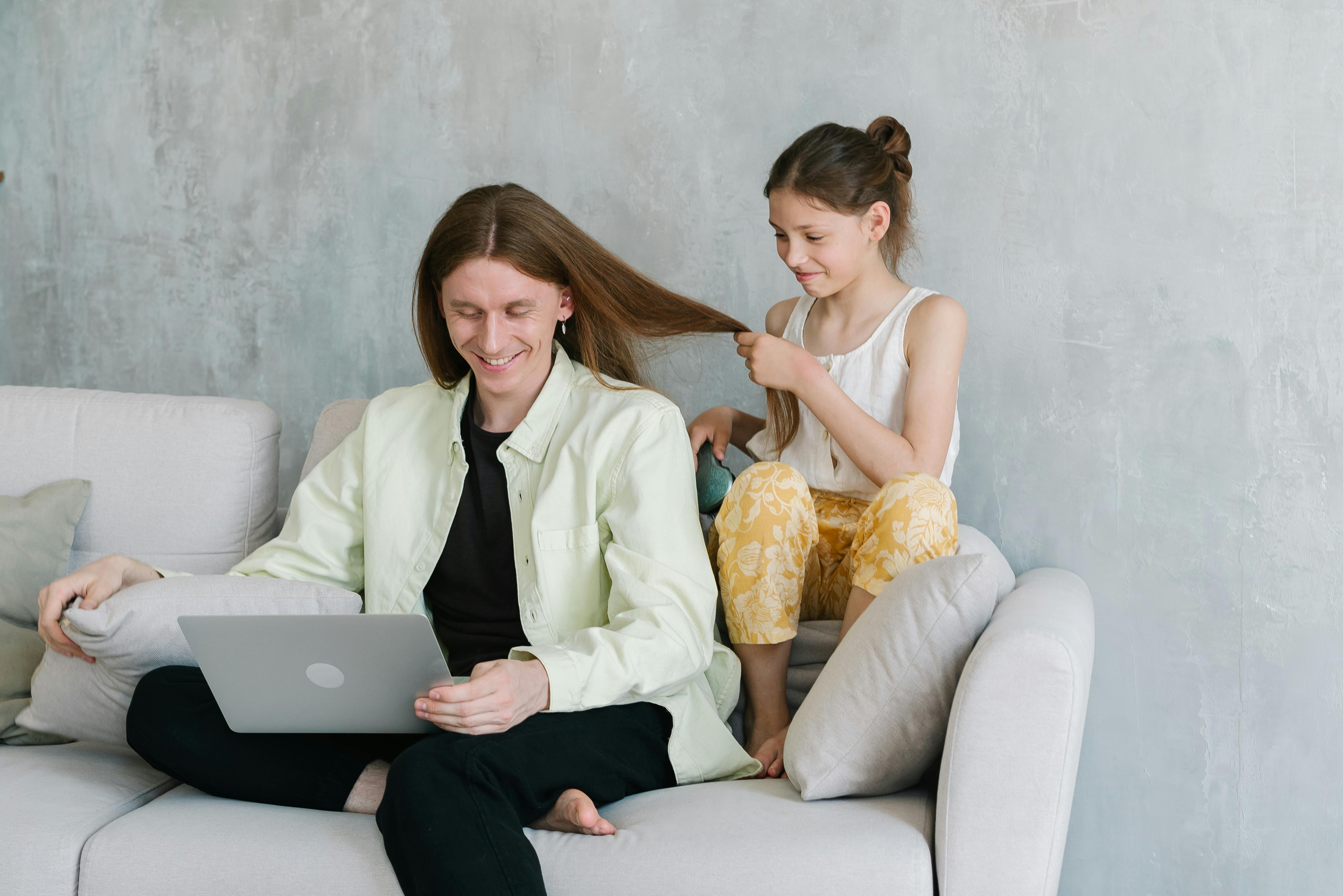 A Young Girl Sitting on the Couch while Holding Her Father's Hair Looking at the Laptop