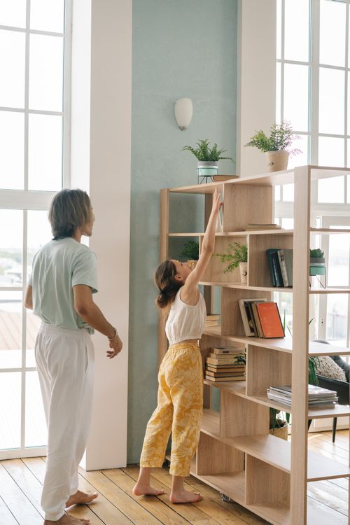 Free A father and daughter bonding while reaching for books on a shelf in a cozy, well-lit living room. Stock Photo