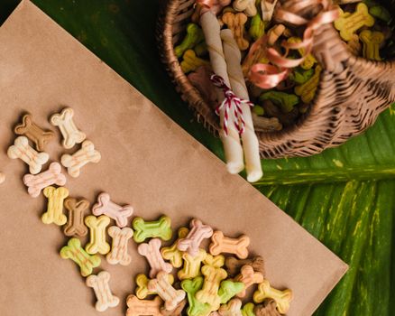 A flat lay of colorful dog bone treats scattered from a woven basket on a leaf surface.