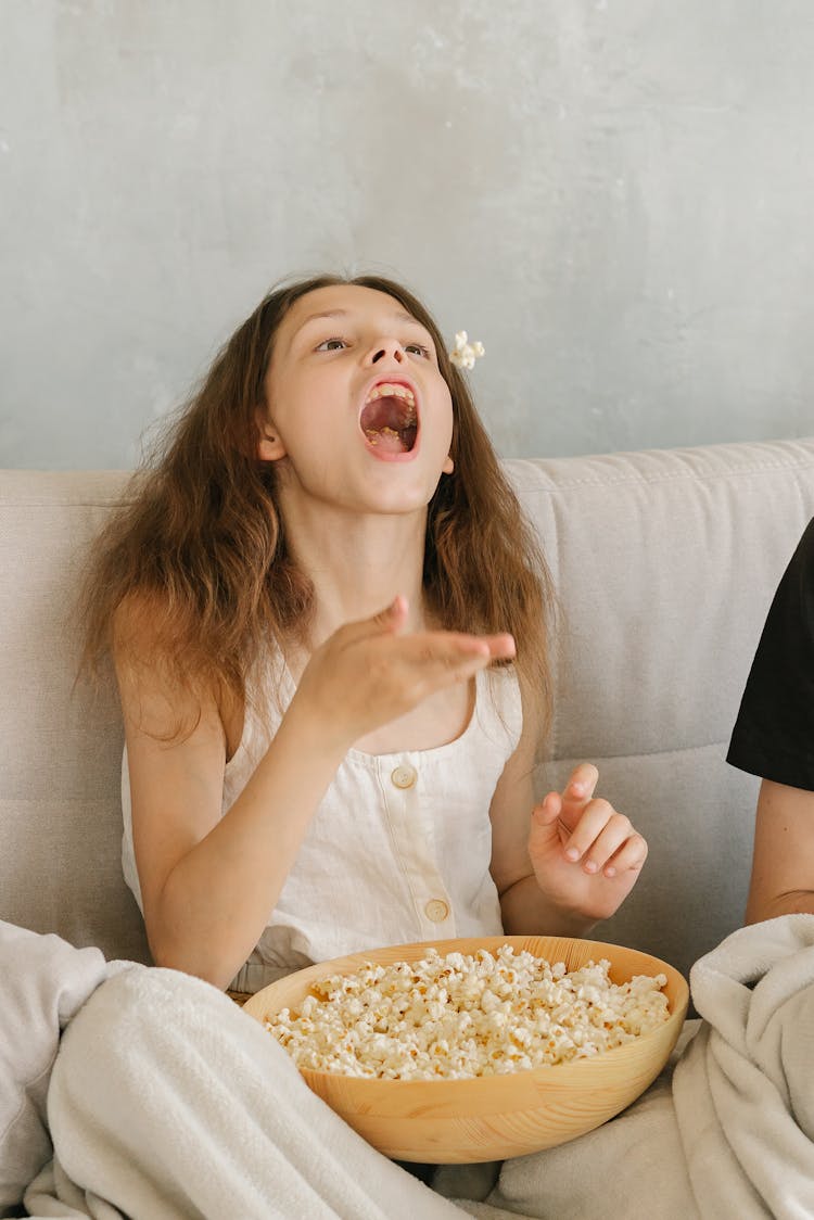 A Young Girl In White Tank Top Eating Popcorn While Sitting On The Couch