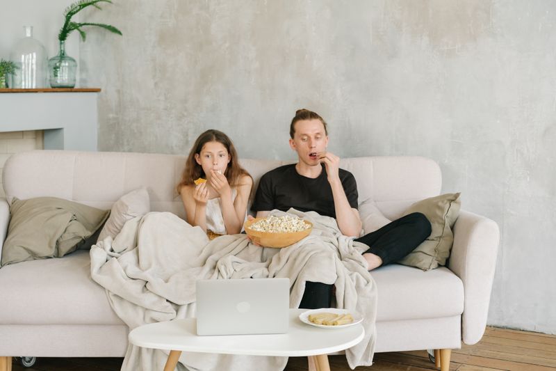 Father and daughter enjoying a cozy movie night with popcorn and snacks on a living room couch.