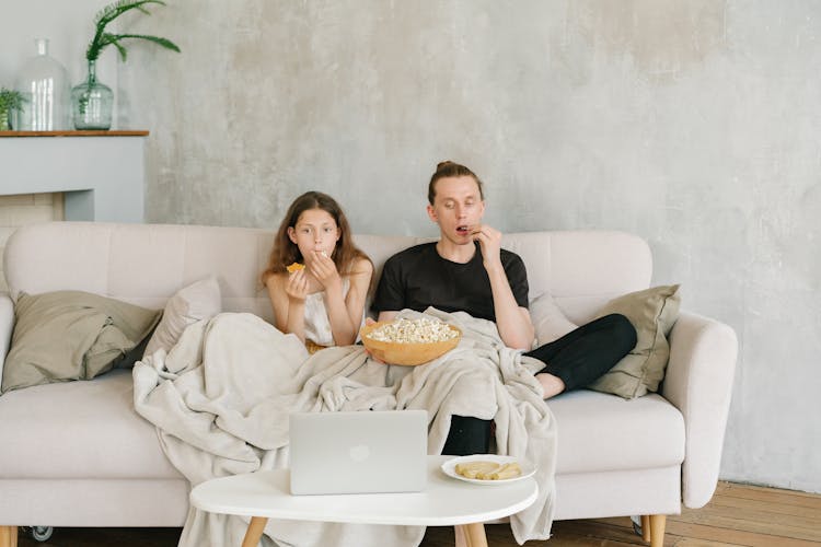 A Father And Daughter Eating Popcorn While Watching On Laptop