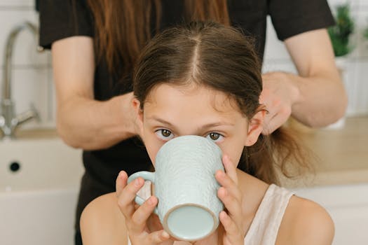 Young girl drinking from a light blue ceramic mug while having her hair styled indoors.