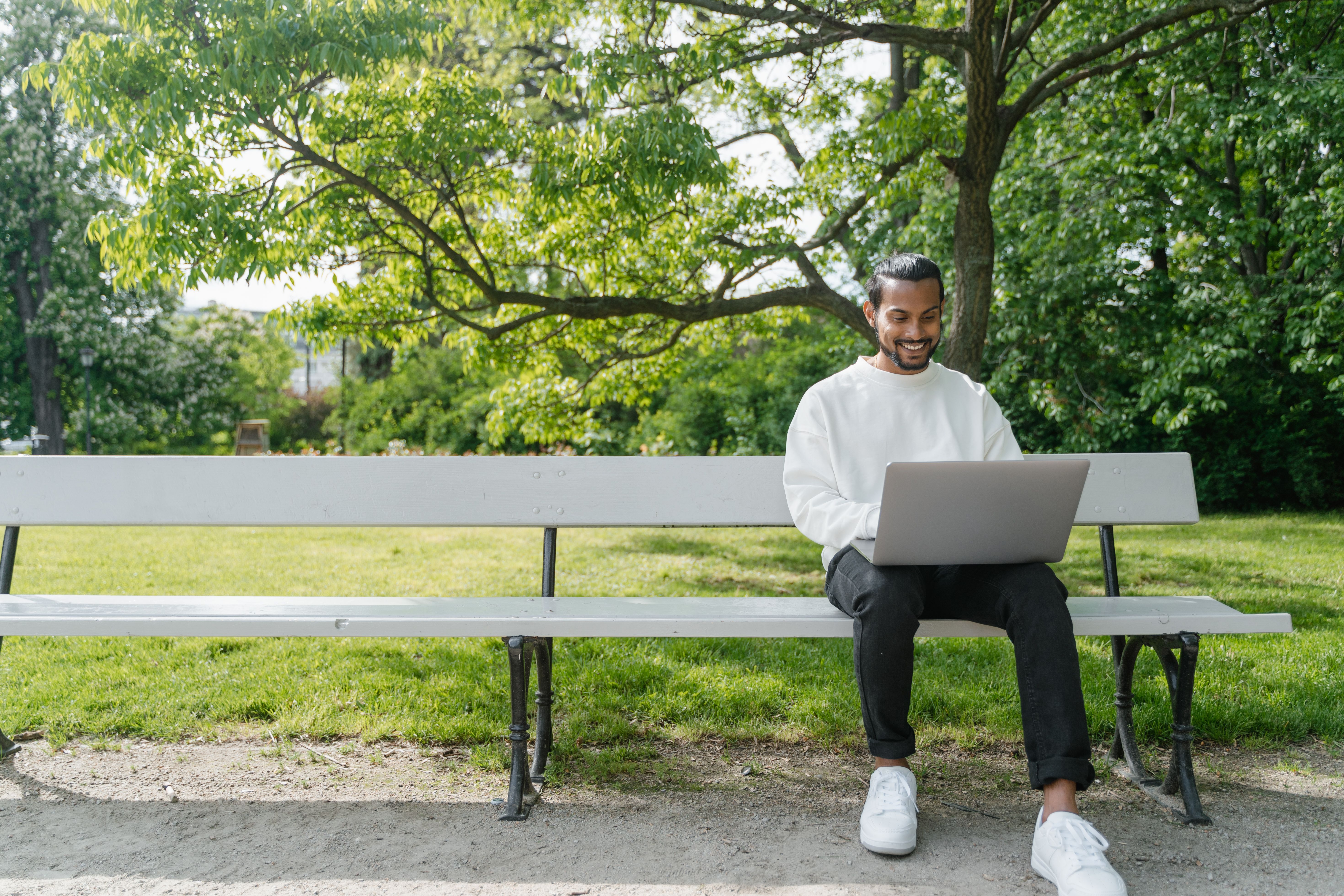 Smiling man in sweater using a laptop while sitting on a park bench surrounded by greenery.