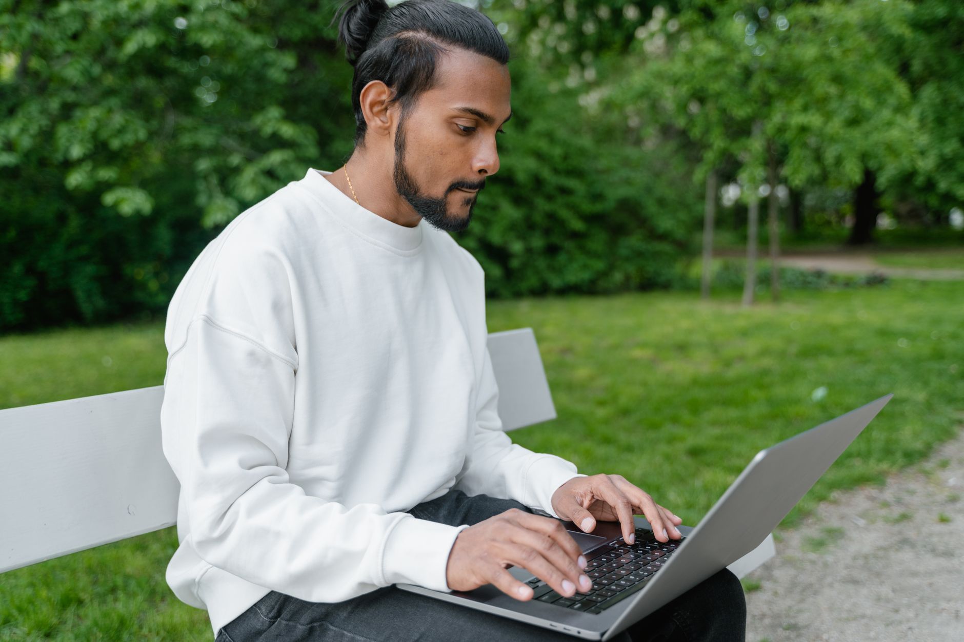 Man in white sweater working on a laptop outdoors in a green park.