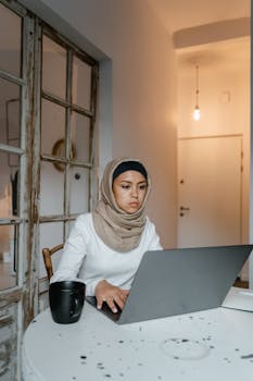 Woman in hijab working on a laptop at a cozy indoor workspace.