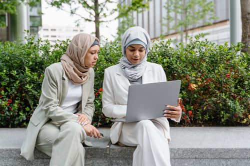 Free Two women in hijabs, discussing work on a laptop, outdoors on a sunny day. Stock Photo