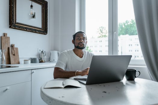 A man in a white t-shirt working on a laptop at a kitchen table, indoors.