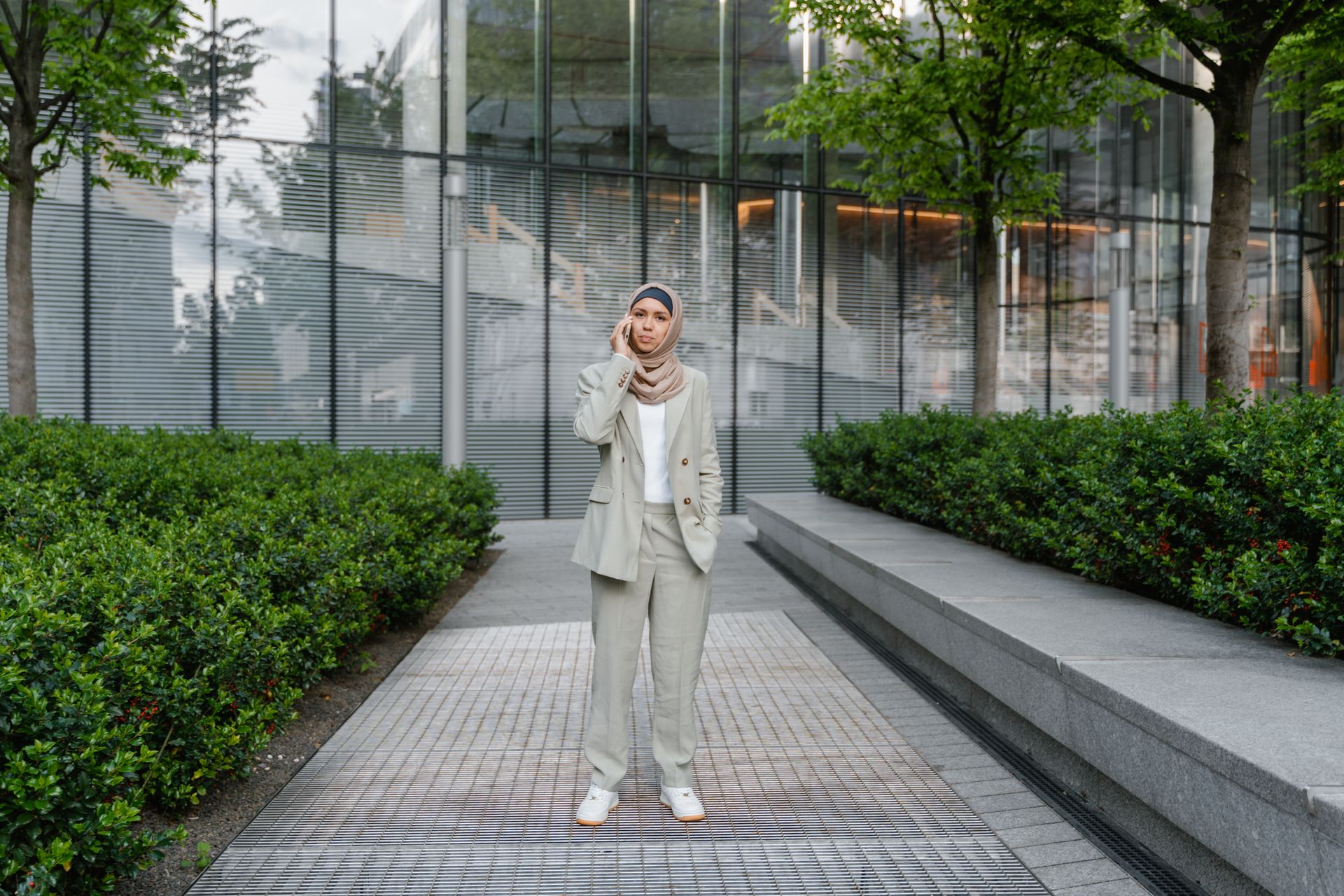 Muslim woman in business attire talks on mobile phone in park with glass building backdrop.