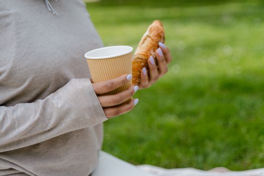 Close-up of hands holding a coffee cup and croissant outdoors on a grassy field.. close, hands, holding