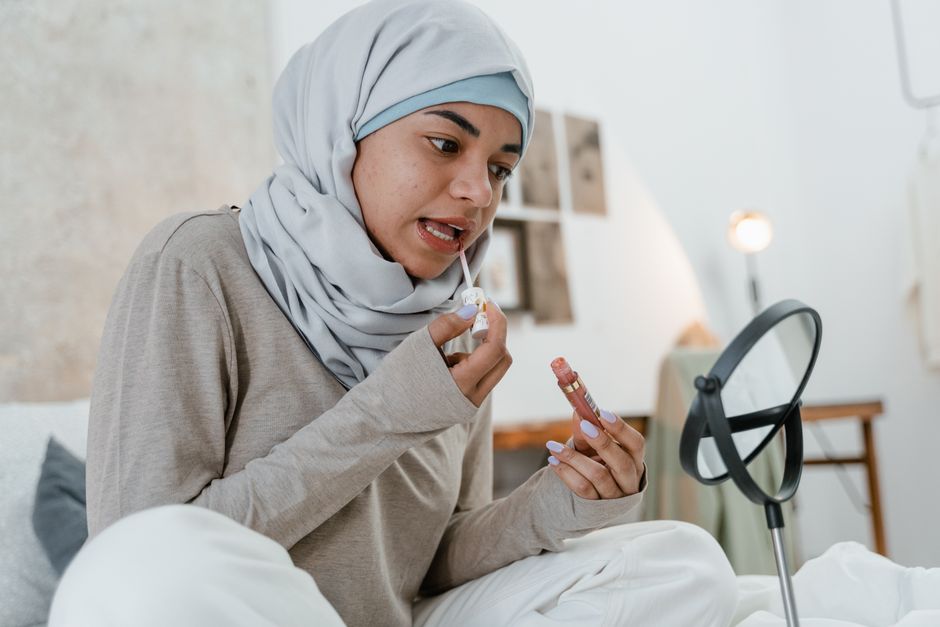 Young Muslim woman applying lipstick while sitting in a cozy bedroom setting with mirror.