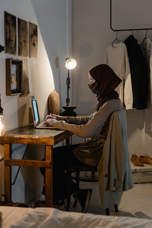 Free Muslim woman in hijab using a laptop at home, sitting by a wooden desk with warm lighting. Stock Photo