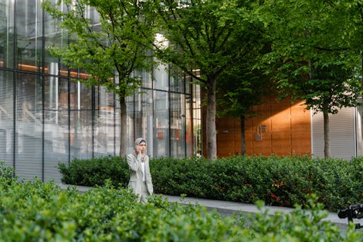 Serious Muslim female in hijab wearing elegant wear having phone conversation while walking near modern building along green shrubs on street