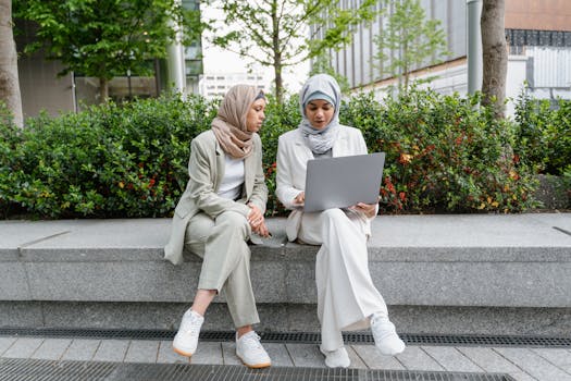 Two Muslim women in business attire working on a laptop outdoors, showcasing modern collaboration.