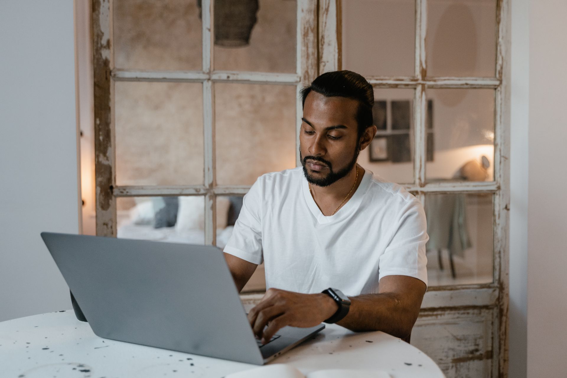 Adult man with beard focused on laptop work at home desk during the day.