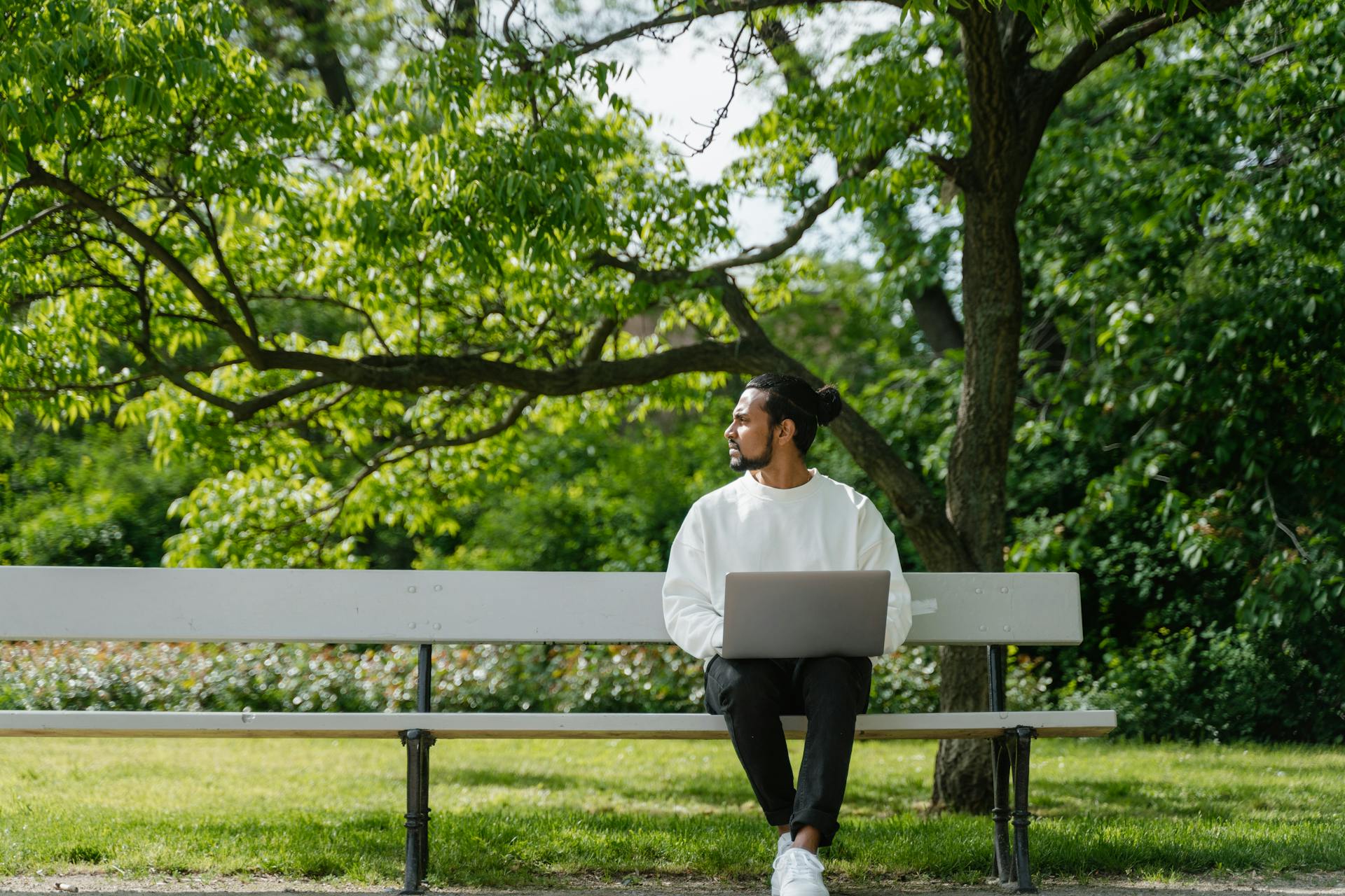 Man in a white sweater working remotely on a laptop while sitting on a park bench.