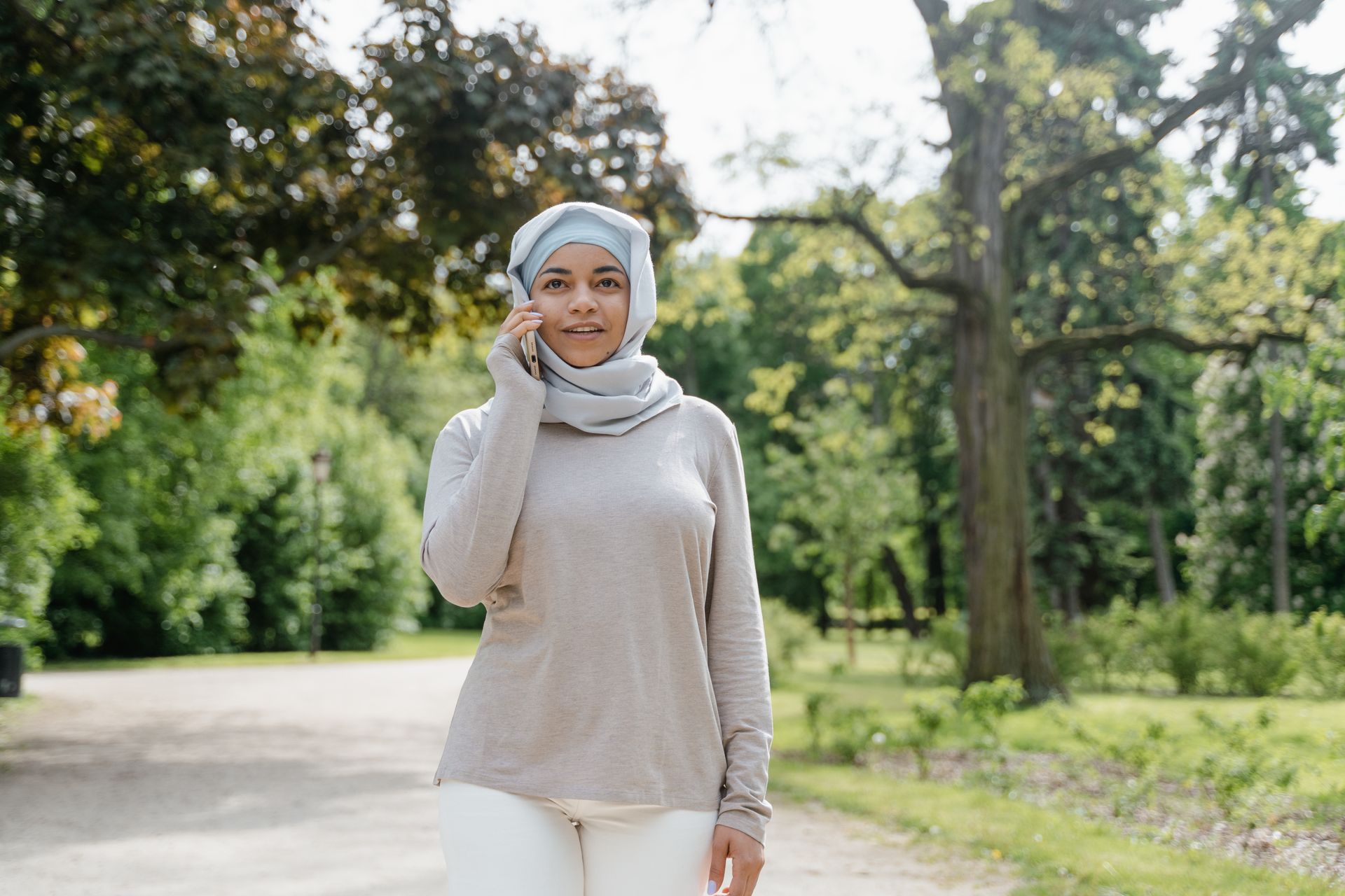 Smiling woman in hijab talking on phone while walking in a sunny park.