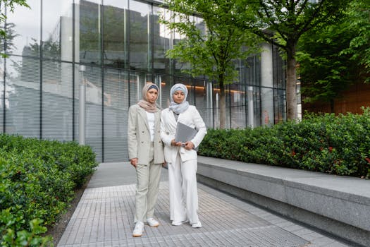 Two Muslim women in business attire, holding a laptop, outdoors in a corporate setting.