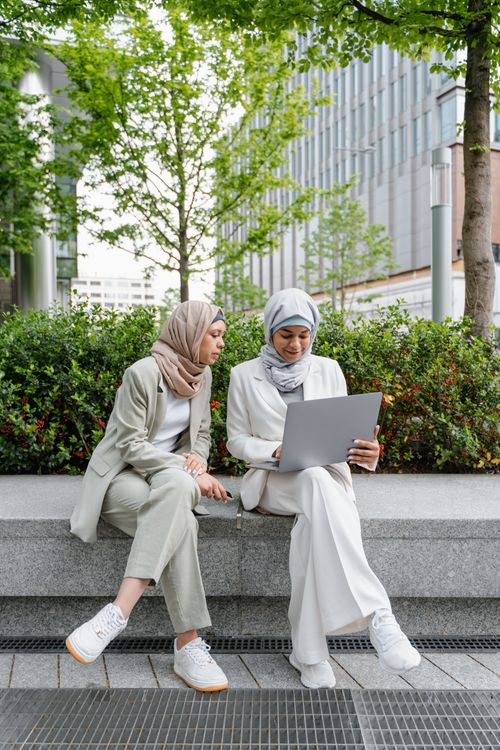 Free Two women in hijabs sitting outdoors on a bench, using a laptop amidst greenery. Stock Photo