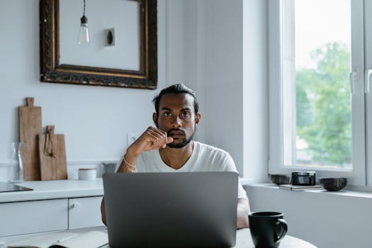 A thoughtful man sits at a desk working on a laptop in a bright, minimalist home setting.