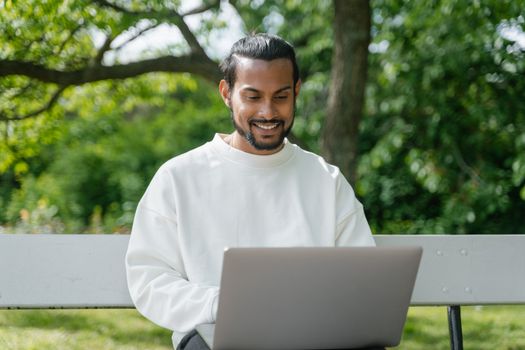 A cheerful man with a beard uses a laptop while sitting on a bench in a lush green park.