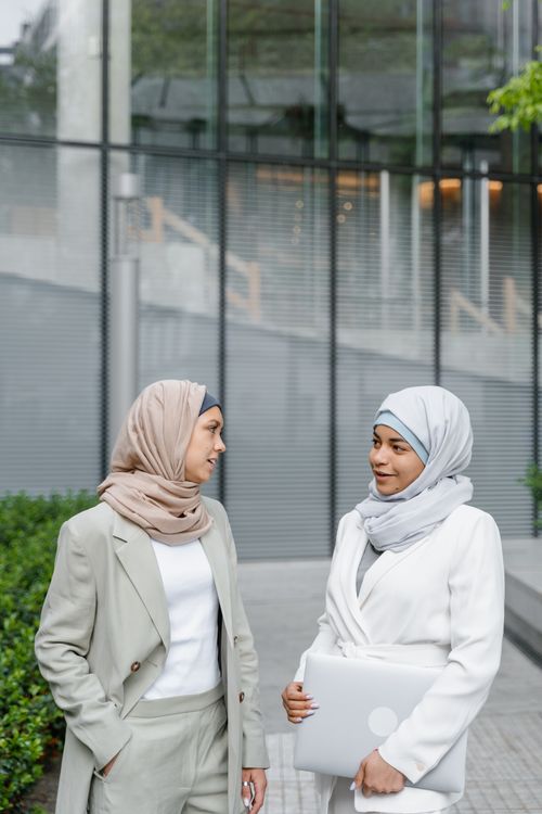 Free Two women in headscarves talking outside a modern building, holding a laptop. Stock Photo