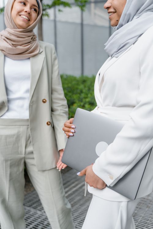 Free Two women wearing hijabs standing outdoors talking while holding a laptop. Stock Photo