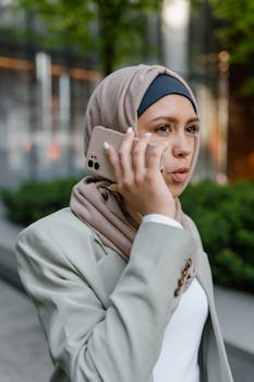 A young woman wearing a hijab is speaking on her smartphone outdoors, dressed in a blazer.