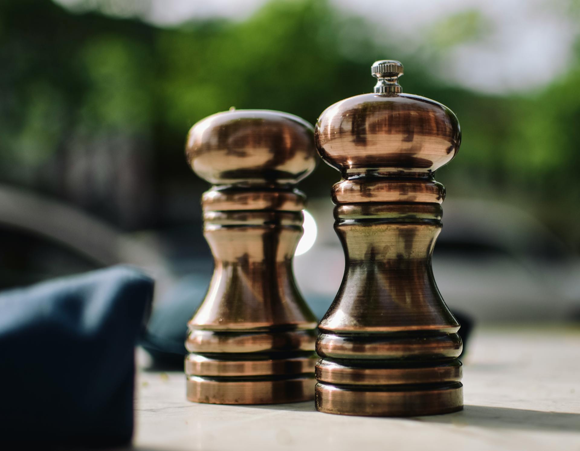 Close-up of copper pepper mill shakers outdoors with green blurred background.