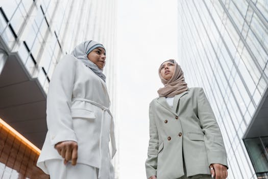 Two professional women in hijabs stand confidently below towering skyscrapers, symbolizing cultural diversity and modern business.